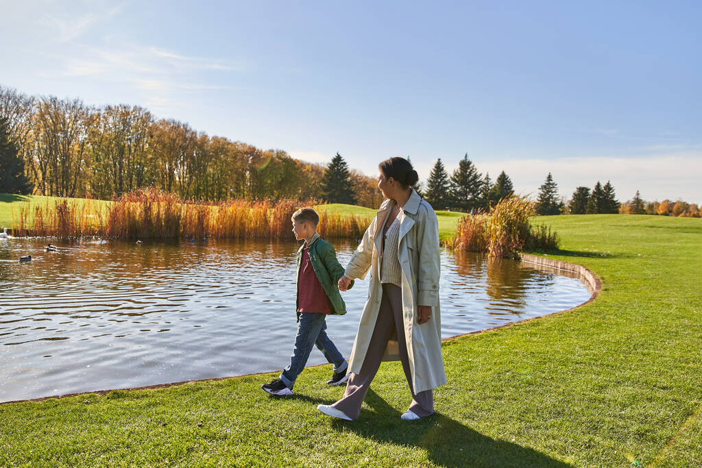 unión familiar, madre afroamericana caminando con su hijo a lo largo del estanque, cogidas de la mano, otoño, naturaleza - Foto, Imagen