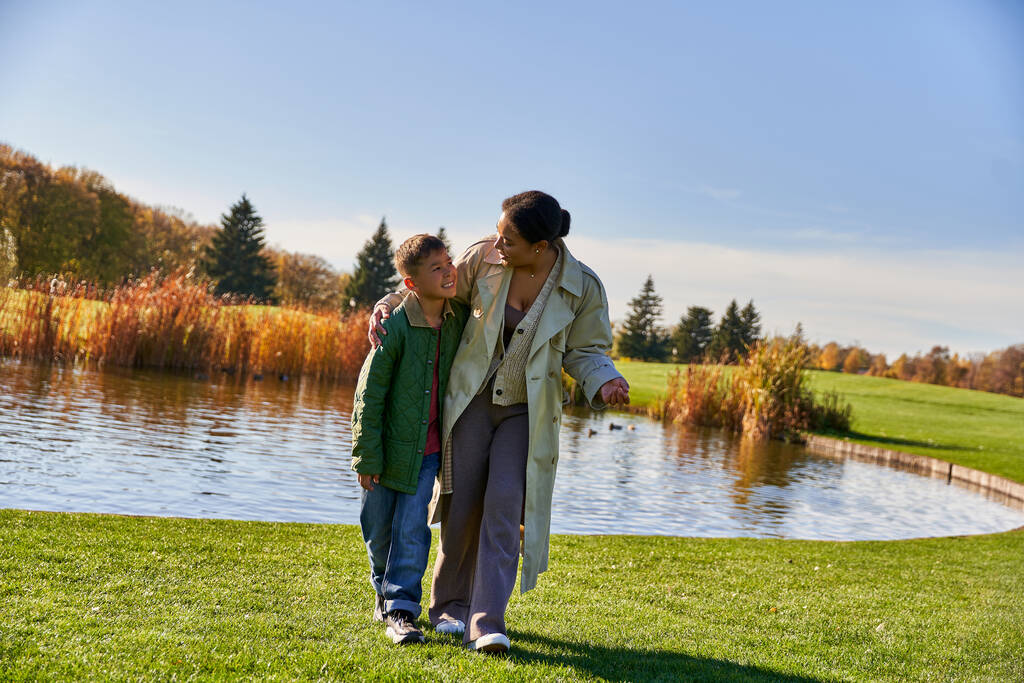 rodinné sbližování, usmívající se africká americká matka procházky se synem podél rybníka, objímání, podzimní sezóna - Fotografie, Obrázek