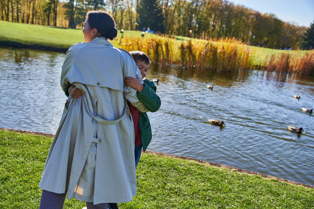 feliz afroamericano madre abrazando hijo cerca de estanque con patos, juguetón, temporada de otoño, alegría, familia - Foto, imagen
