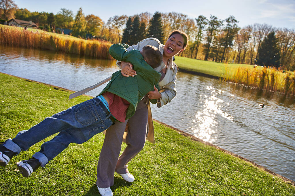 playful, joy, happy african american mother lifting son, having fun near pond, outerwear, autumn - Photo, Image