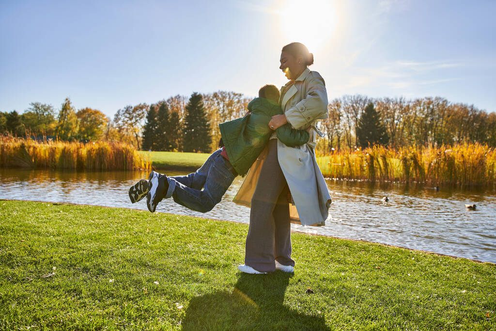 відвертий, щасливий афроамериканська мати піднімає грайливого сина, розважається біля ставка, верхній одяг, осінь - Фото, зображення