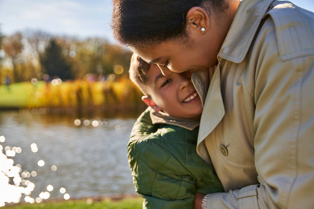 candid, autumn season, happy african american woman in outerwear hugging cheerful son, portrait - Photo, Image