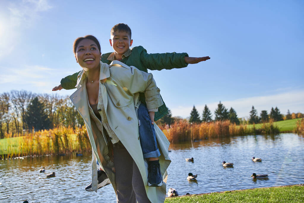 joyful mother piggybacking son near pond with ducks, childhood, african american, autumn, free - Photo, Image
