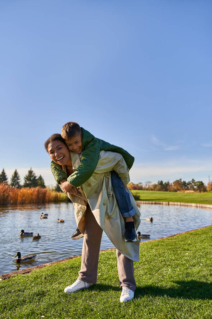 colores de otoño, alegre afroamericana mujer piggybacking chico cerca de estanque con patos, infancia, otoño - Foto, Imagen