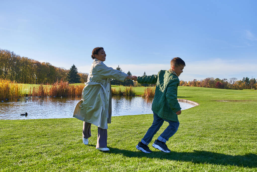playful african american mother and son running on grass near pond, modern parenting, fall fashion - Photo, Image