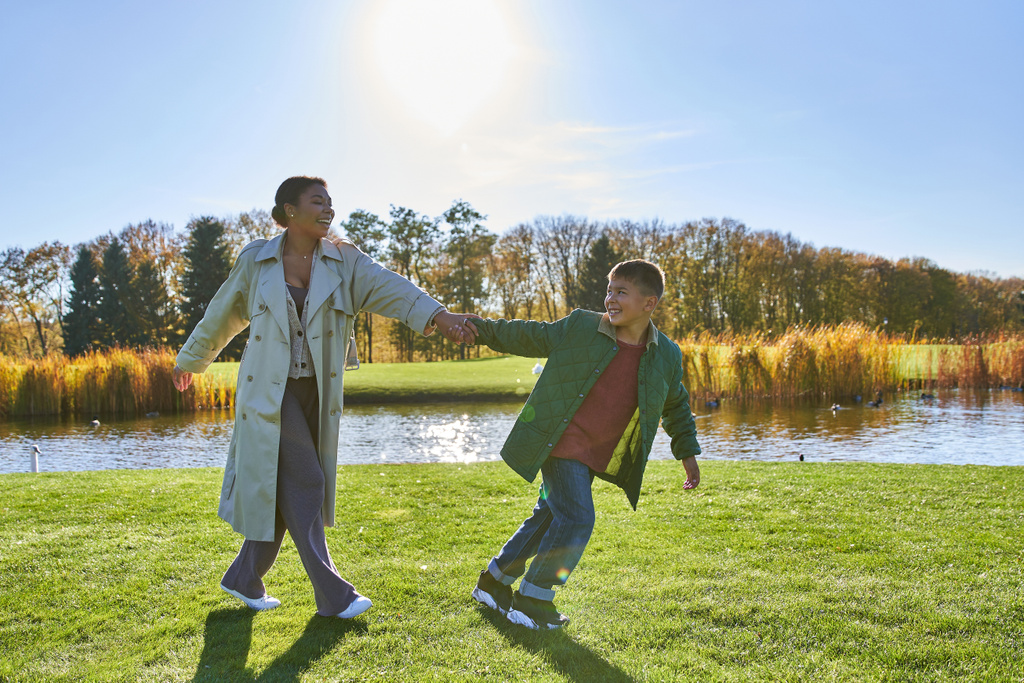 息子に続く陽気なアフリカ系アメリカ人の母親,池の近くを歩く,近代的な子育て,秋のファッション - 写真・画像