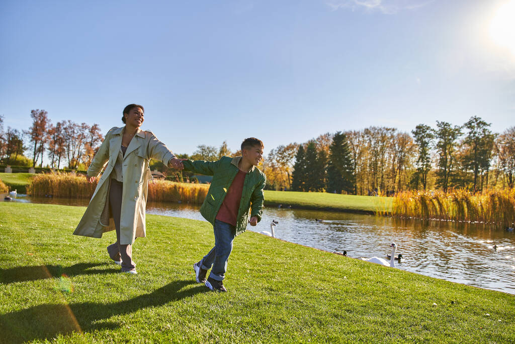 happy childhood, african american woman in outerwear running with son near pond, autumn, fall season - Photo, Image