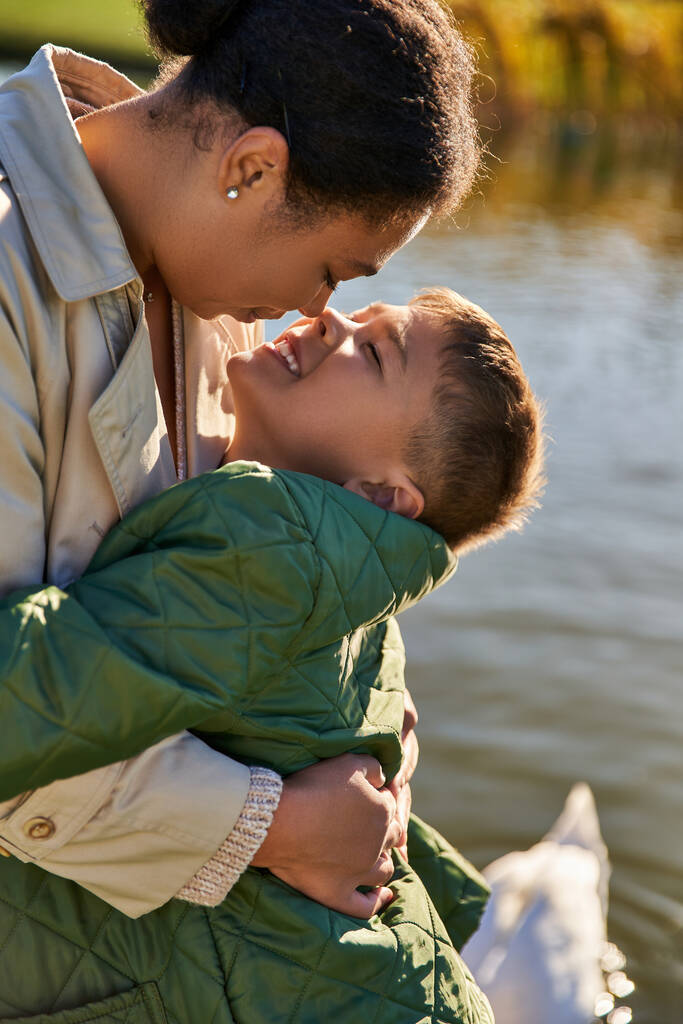 amor maternal, niño feliz abrazando a la madre cerca del lago, familia afroamericana, temporada de otoño, otoño - Foto, imagen