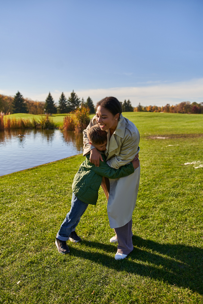 happy woman hugging son, standing near lake in park, african american family, autumn fashion - Photo, Image