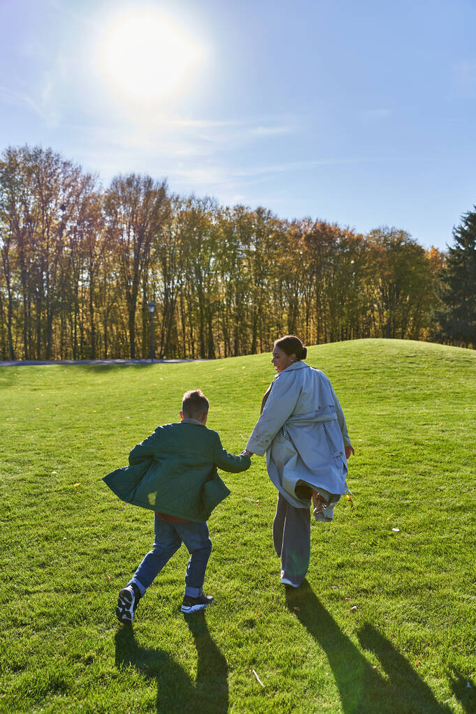 día soleado en otoño, mujer afroamericana corriendo junto con su hijo en el parque, hierba, moda otoñal - Foto, Imagen