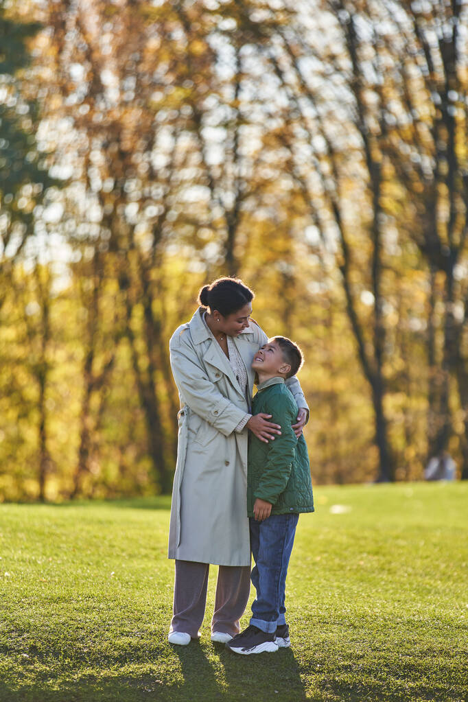 veselý africký americký maminka a syn objímání v podzimním parku, žena a chlapec v outterwear - Fotografie, Obrázek