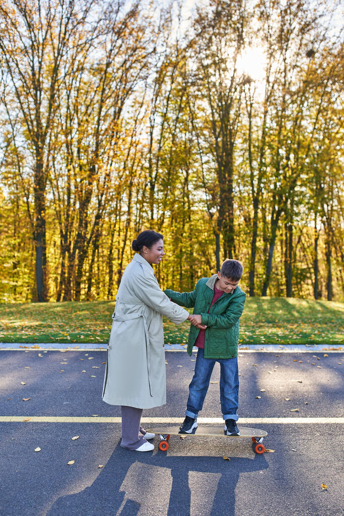 moeder en zoon in park, gelukkig Afrikaans amerikaans vrouw hand in hand met jongen op penny board, herfst - Foto, afbeelding