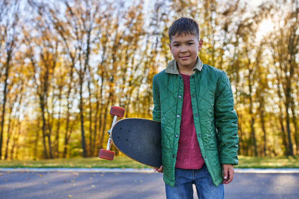feliz afroamericano chico en ropa de abrigo celebración penny board, parque de otoño, temporada de otoño, retrato - Foto, Imagen