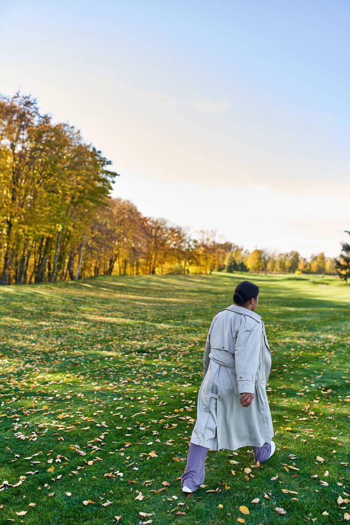 achteraanzicht van Afrikaanse Amerikaanse vrouw in trench jas lopen op gras met gouden bladeren, herfst park - Foto, afbeelding