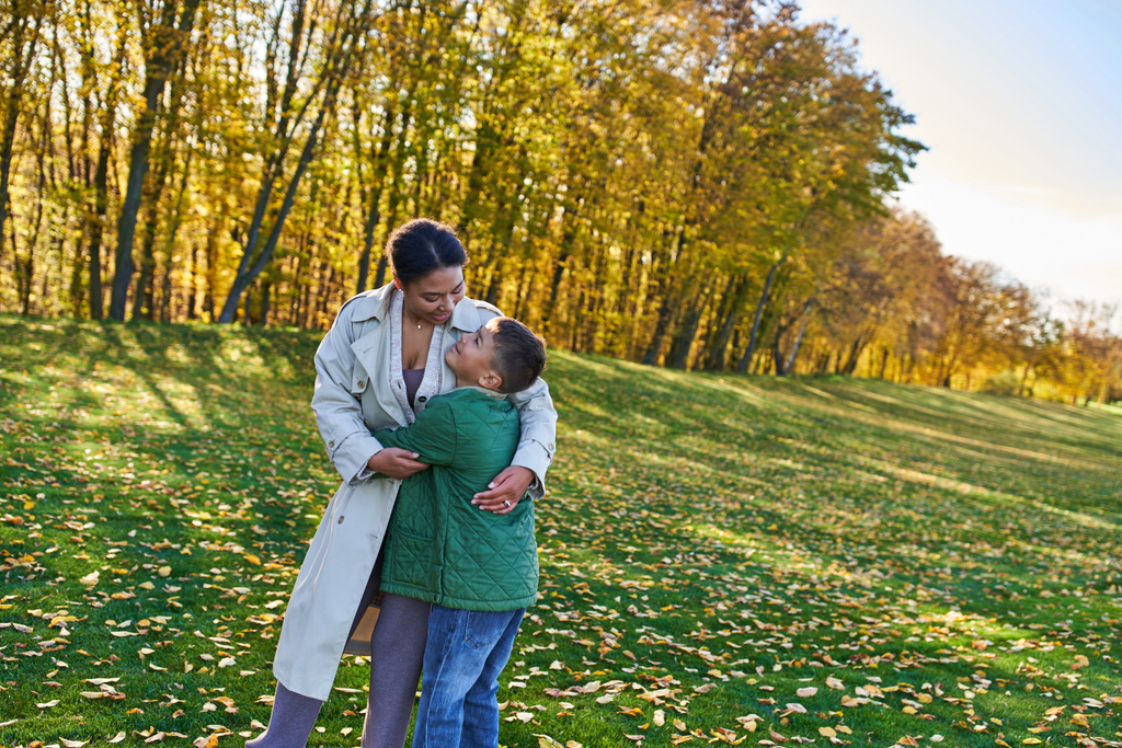 szczęśliwa Afroamerykanka przytulająca się do syna, stojąca na trawie ze złotymi liśćmi, jesień - Zdjęcie, obraz