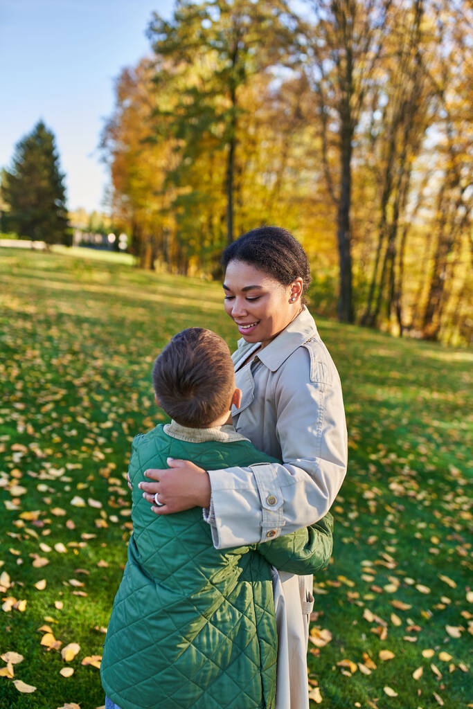 amor maternal, mujer afroamericana feliz mirando a lindo hijo, de pie cerca de hojas doradas, otoño - Foto, Imagen