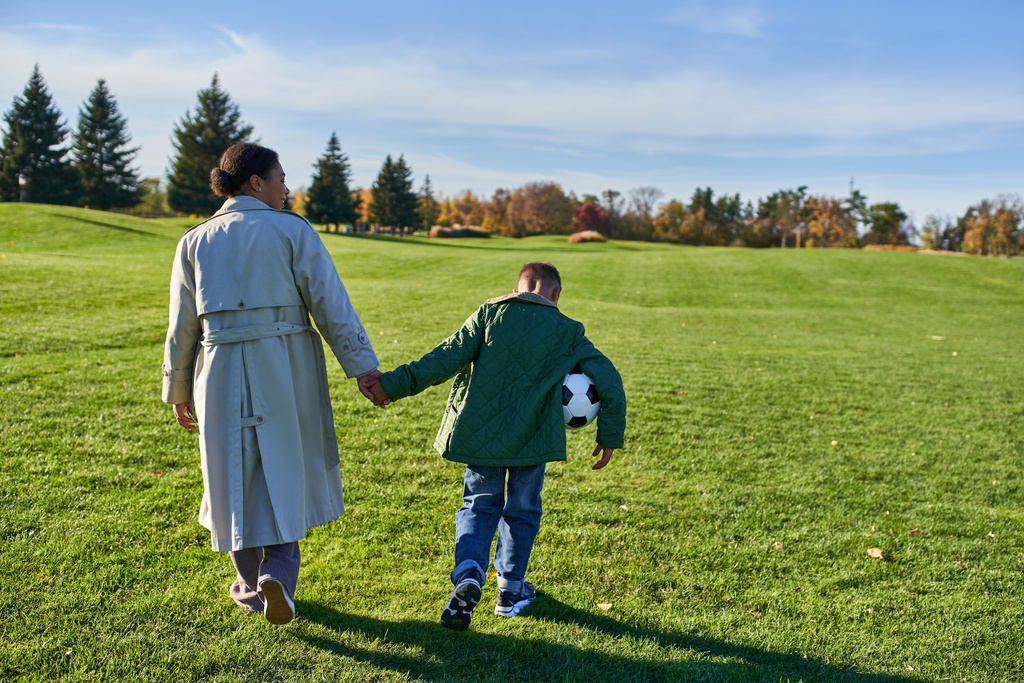 ragazzo afroamericano che tiene il calcio, cammina con la mamma sul campo verde, si tiene per mano, autunno - Foto, immagini
