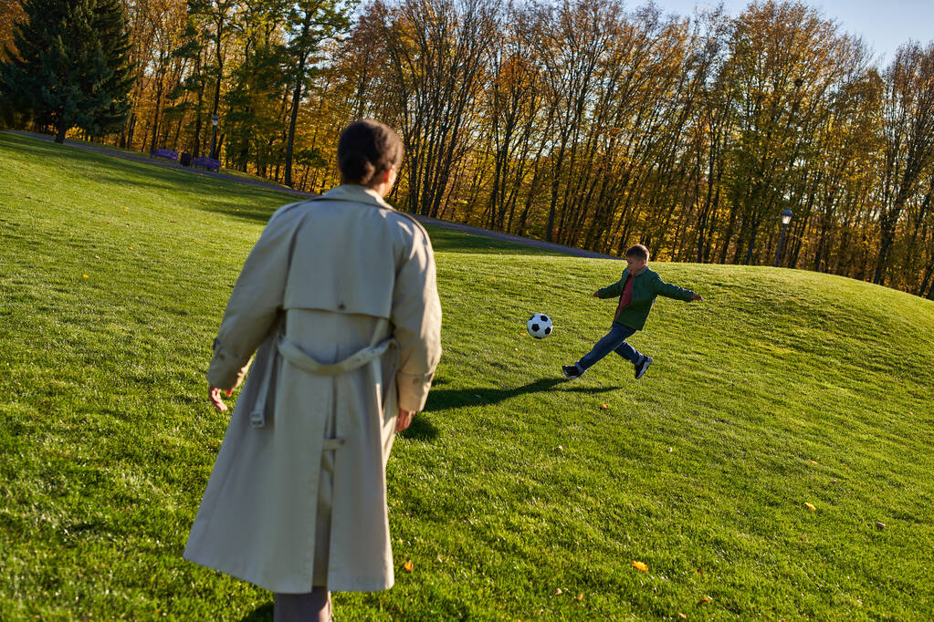 χαριτωμένο αφροαμερικανό αγόρι που παίζει ποδόσφαιρο κοντά στη μητέρα στο πράσινο πεδίο, ποδόσφαιρο, φθινόπωρο, φθινοπωρινή σεζόν - Φωτογραφία, εικόνα