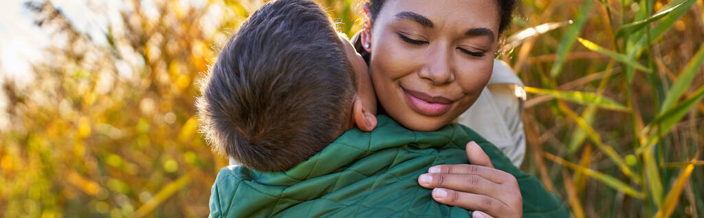 ευτυχία, μητρική αγάπη, αφροαμερικάνικη μητέρα αγκαλιάζει τον γιο της με φθινοπωρινά εξωτερικά ενδύματα, πανό - Φωτογραφία, εικόνα