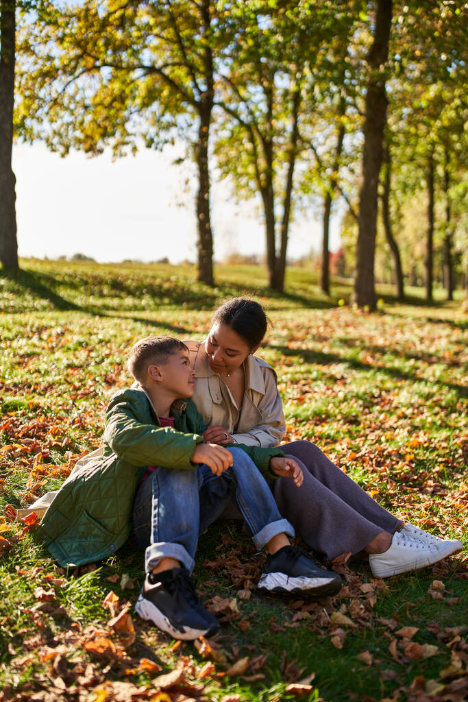 mütterliche Liebe, glückliche afrikanisch-amerikanische Frau und Sohn sitzen auf Gras mit goldenen Blättern, Herbst - Foto, Bild