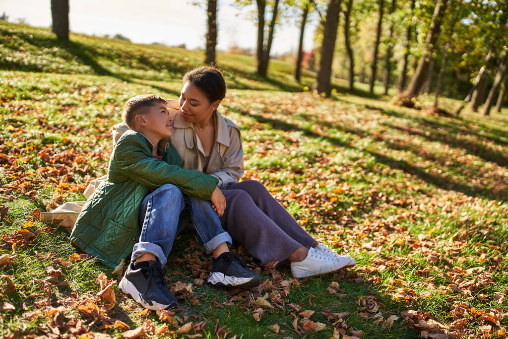 amor maternal, alegre mujer afroamericana e hijo sentado en la hierba con hojas doradas, otoño - Foto, imagen