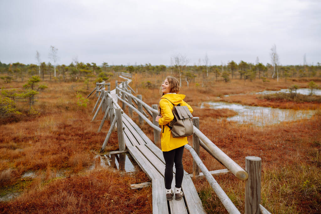 Mujer alegre en un abrigo amarillo camina, explora un camino de madera de los humedales. El concepto de viajes, vacaciones, naturaleza. Estilo de vida activo. - Foto, imagen