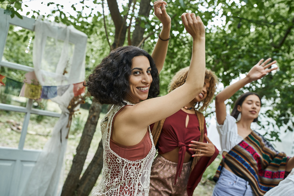positive multicultural woman looking at camera while dancing near friends in retreat center - Photo, Image