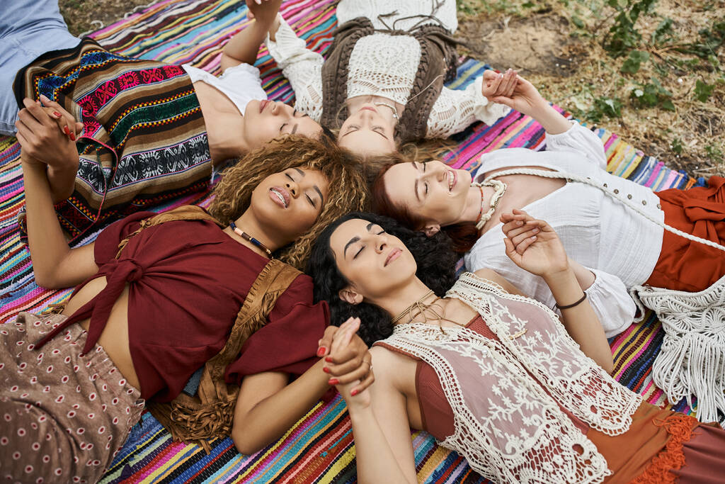 vysoký úhel pohledu na usmívající se multietnické ženy držící se za ruce a ležící na přikrývce v centru ústupu - Fotografie, Obrázek