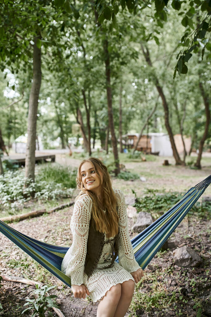 smiling young blonde woman in boho dress sitting in hammock in retreat center - Photo, Image