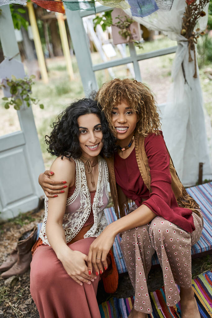 smiling interracial girlfriends in boho outfits hugging and looking at camera in retreat center - Photo, Image