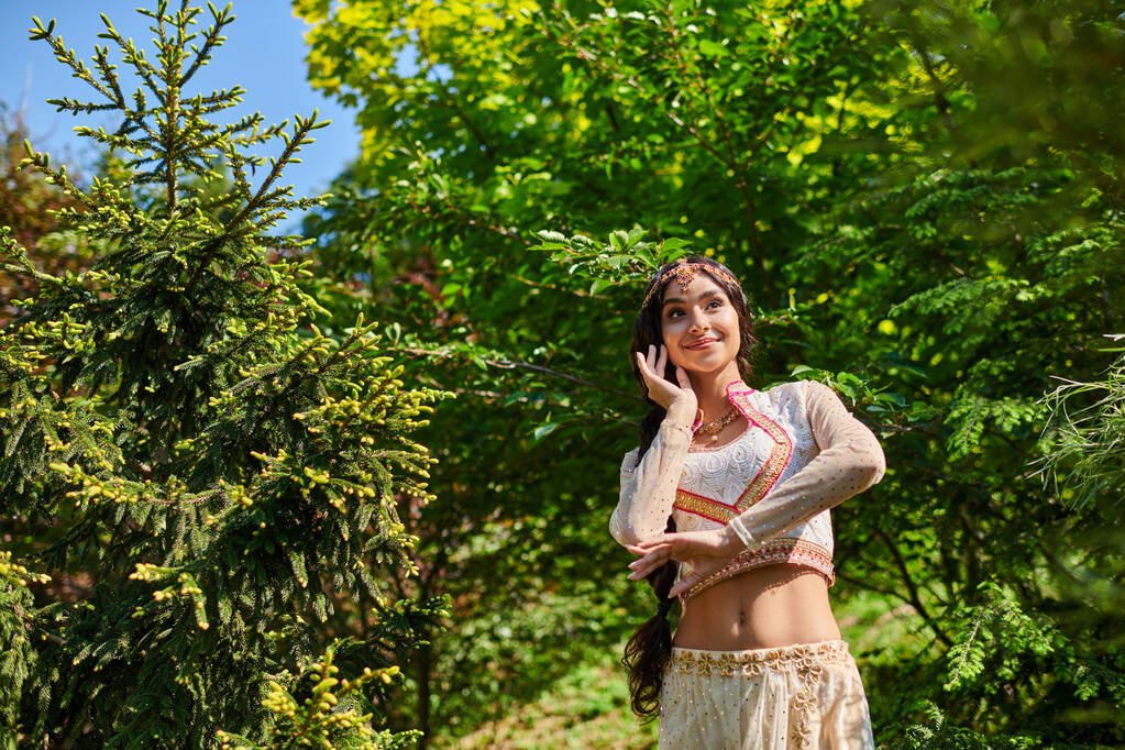 беззаботная индийская женщина в стильной традиционной одежде танцует в летнем парке в солнечный день - Фото, изображение