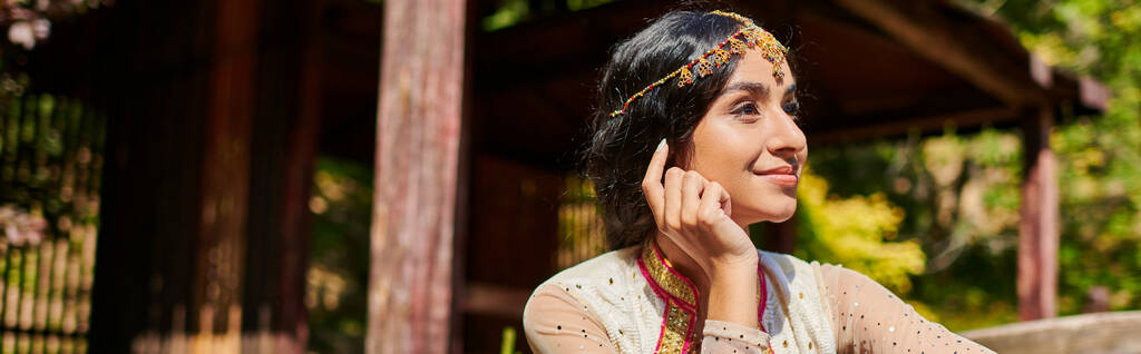 μελαχρινή, χαμογελαστή και ονειρεμένη Ινδή γυναίκα με παραδοσιακά ρούχα που κοιτάζει μακριά στο καλοκαιρινό πάρκο - Φωτογραφία, εικόνα