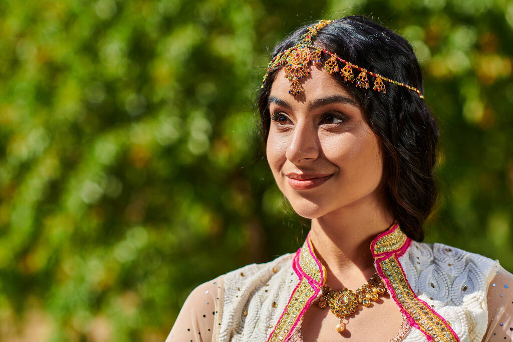 kuva tyytyväinen, brunette intialainen nainen etnisissä vaatteissa katselee pois ja hymyilee kesäpuistossa - Valokuva, kuva