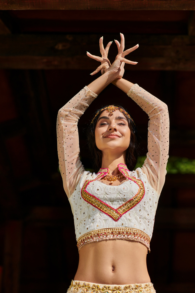 jovem mulher indiana alegre no elegante desgaste étnico dançando com os olhos fechados e mãos levantadas ao ar livre - Foto, Imagem