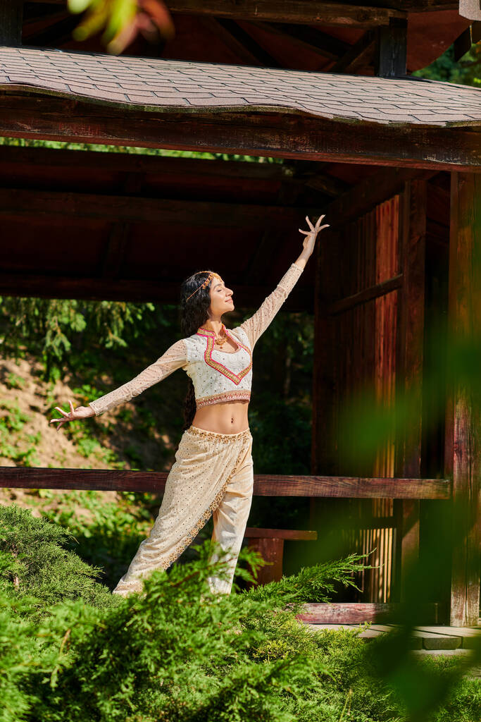 zomer park, indiaanse vrouw in elegante etnische kleding dansen in de buurt van houten nis in de zomer park - Foto, afbeelding