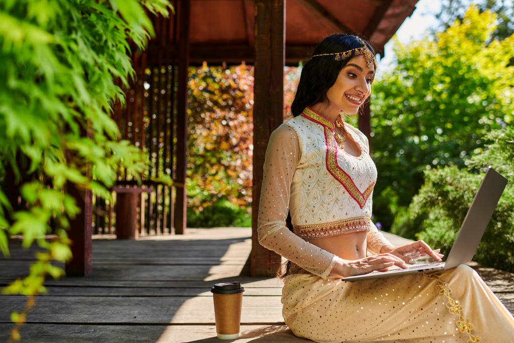 letni park, szczęśliwy etniczny styl kobieta wpisując na laptopie w pobliżu kawy, aby przejść w drewnianej alkowie - Zdjęcie, obraz