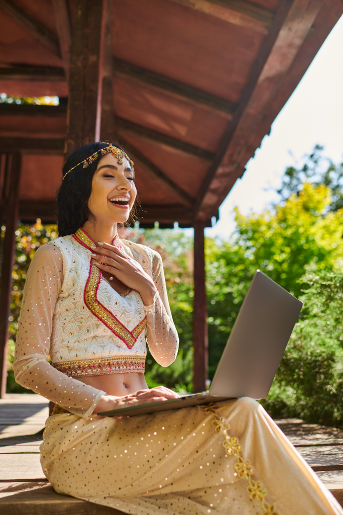 podekscytowana Hinduska kobieta w etnicznym ubraniu patrząca na laptopa i śmiejąca się w drewnianej alkowie letniego parku - Zdjęcie, obraz