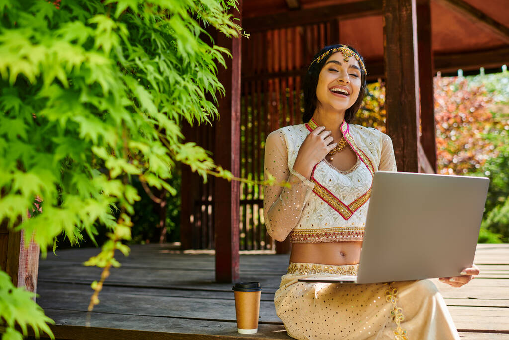 γελώντας ινδή γυναίκα κάθεται με φορητό υπολογιστή κοντά στον καφέ για να πάει στο ξύλινο alcove την ημέρα του καλοκαιριού - Φωτογραφία, εικόνα