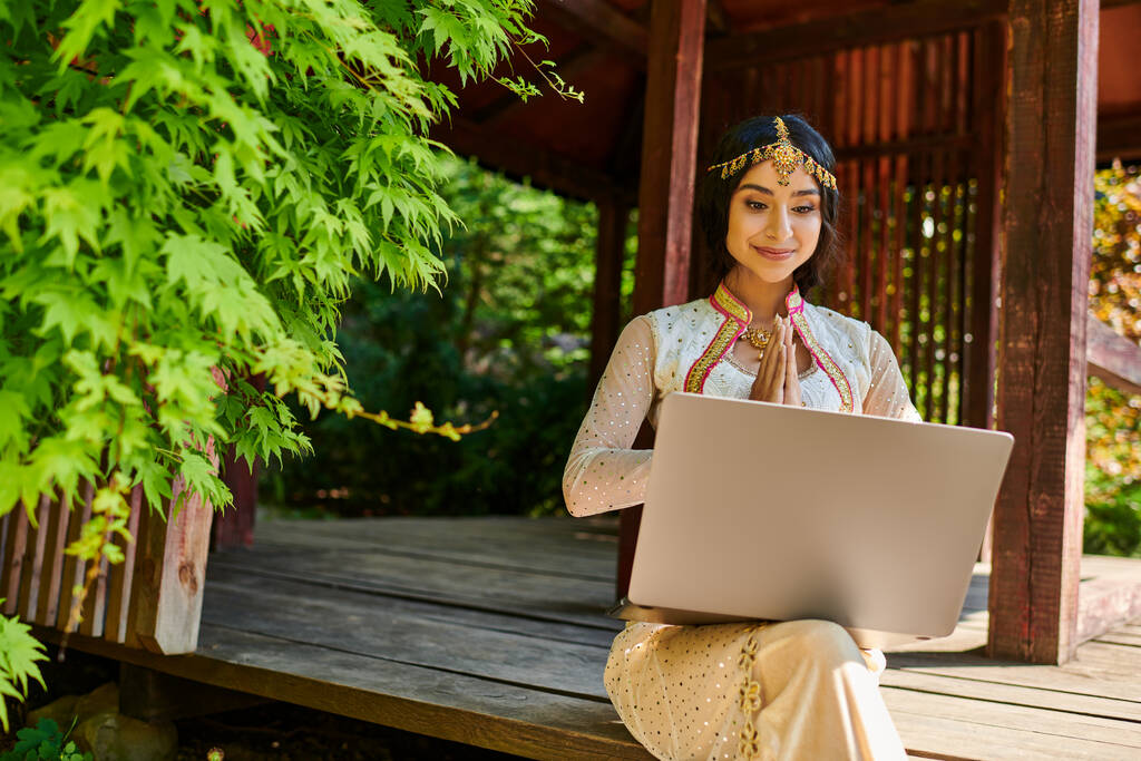 улыбающаяся индианка в традиционной одежде, показывающая приветственный жест во время видеозвонка на ноутбуке - Фото, изображение