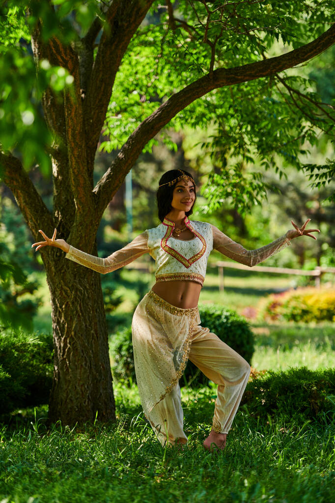 витончена індійська жінка в яскравому етнічному вбранні танцює під деревом в літньому парку, щастя - Фото, зображення