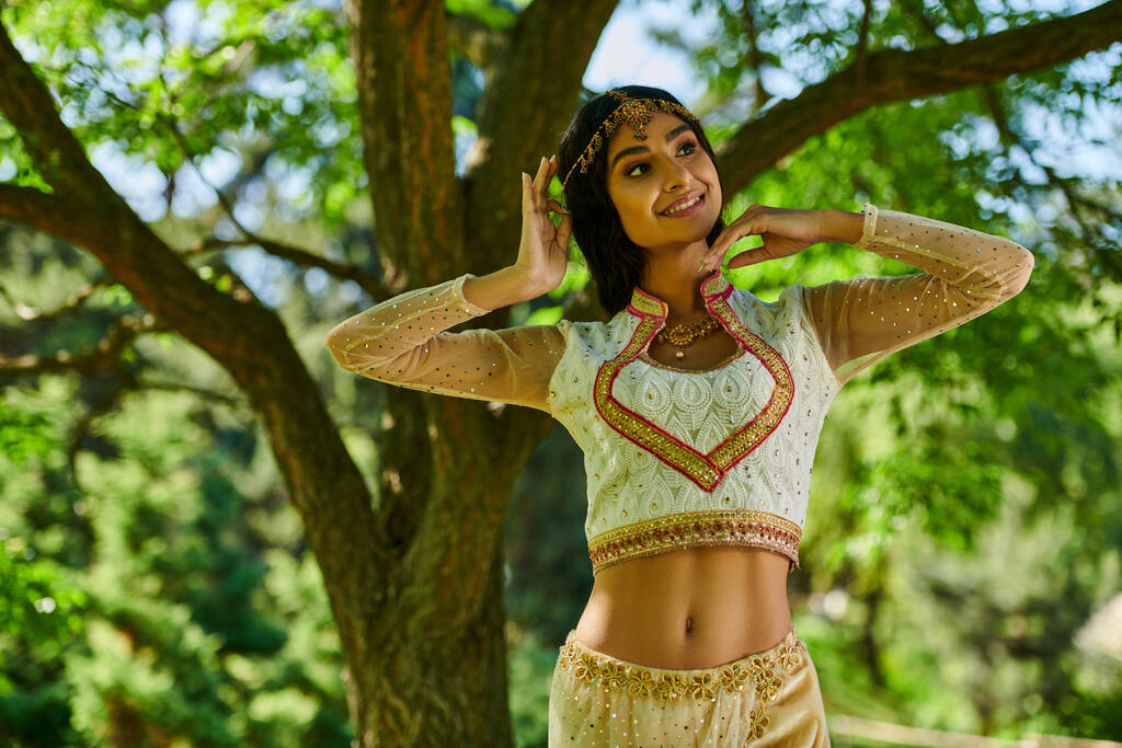 mujer india elegante en ropa tradicional sonriendo y mirando hacia otro lado en el parque verde, ocio de verano - Foto, imagen