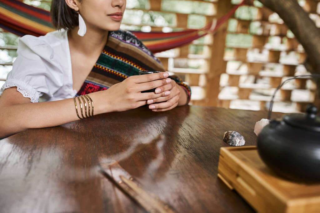 vista cortada de jovem mulher em estilo boho roupas sentadas com xícara de chá na mesa de madeira - Foto, Imagem