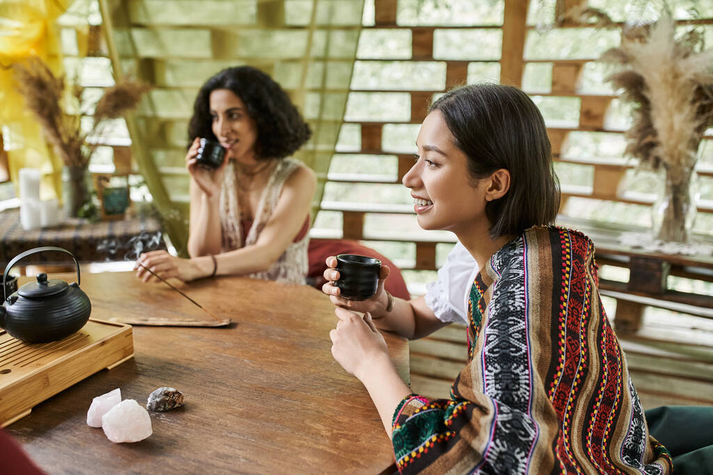 veselá mladá žena s šálkem čaje v blízkosti multiraciální přítelkyně u dřevěného stolu v centru ústupu - Fotografie, Obrázek