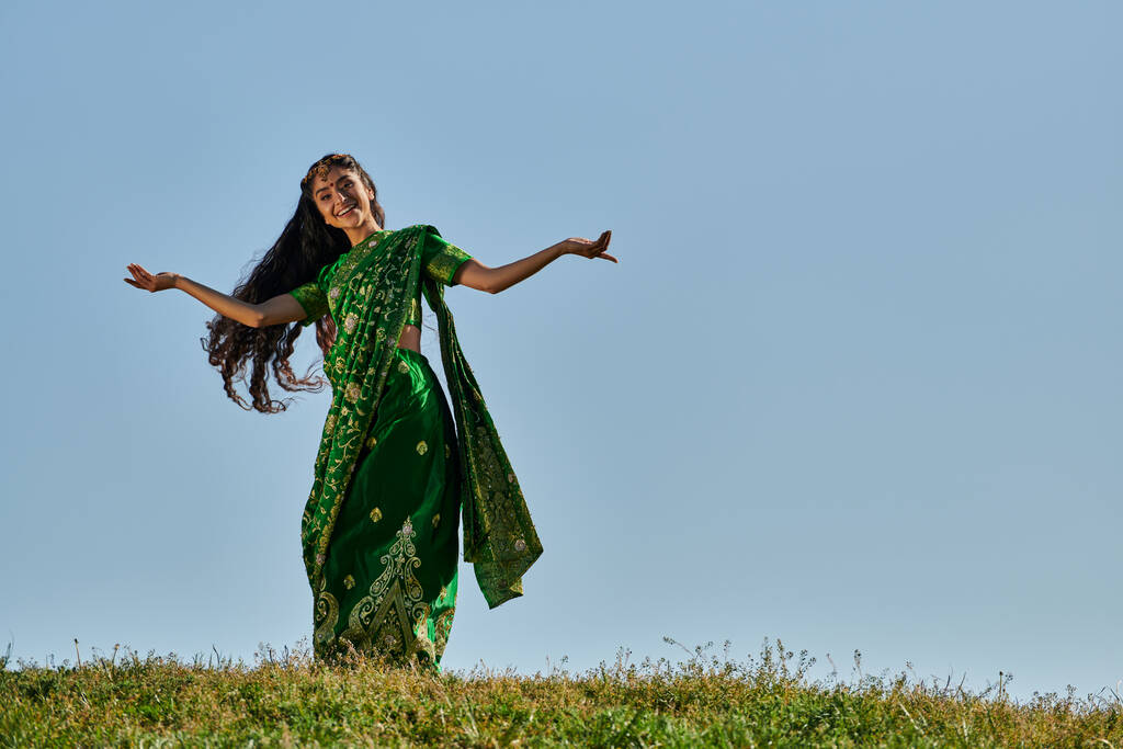 carefree indian woman in traditional sari smiling at camera on green lawn under blue sky - Photo, Image