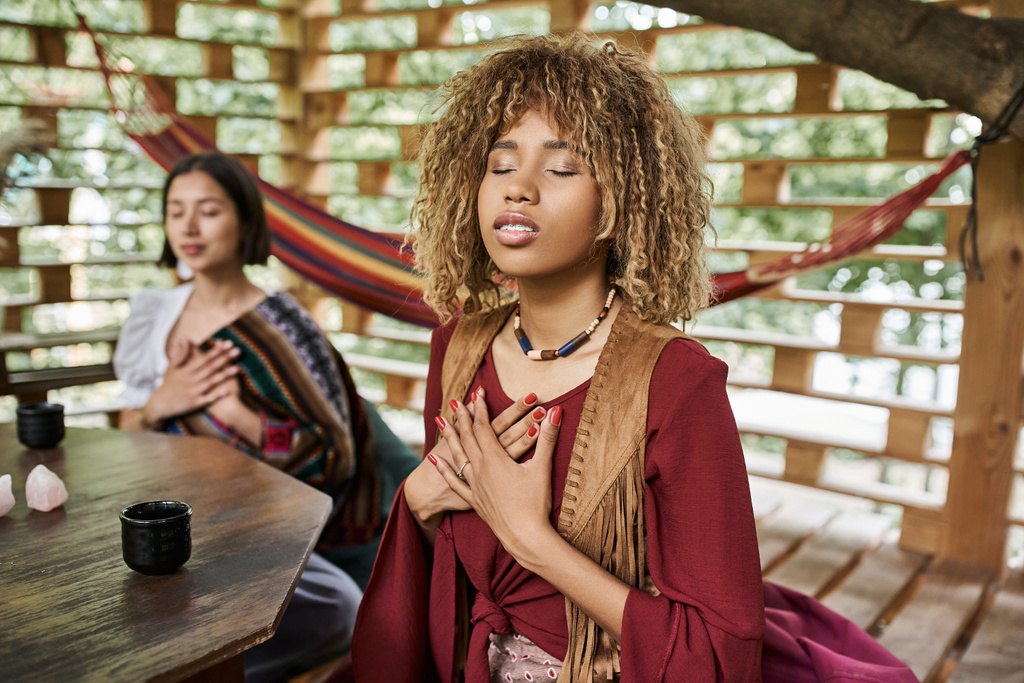 Afričanky americká žena v boho stylu oblečení meditace se zavřenýma očima v blízkosti rozmazané přítelkyně - Fotografie, Obrázek