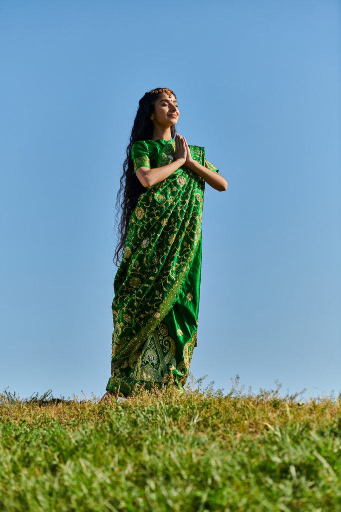 felice donna indiana in sari con le mani in preghiera e gli occhi chiusi sul prato sotto il cielo blu, giorno d'estate - Foto, immagini