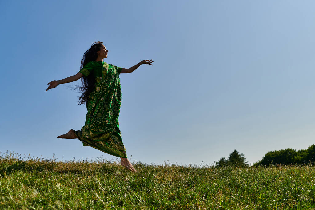wolnomyślicielka indyjska w sari biegająca po zielonej łące pod błękitnym niebem, szczęśliwe lato - Zdjęcie, obraz