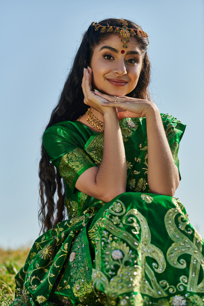 sonriente mujer india en sari y matha patti mirando la cámara en la colina cubierta de hierba y el cielo en el fondo - Foto, Imagen