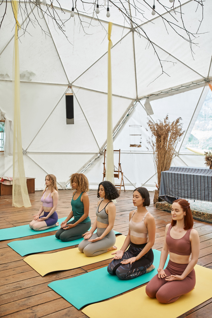 multiracial girlfriends in sportswear meditating in thunderbolt pose in modern retreat center - Photo, Image