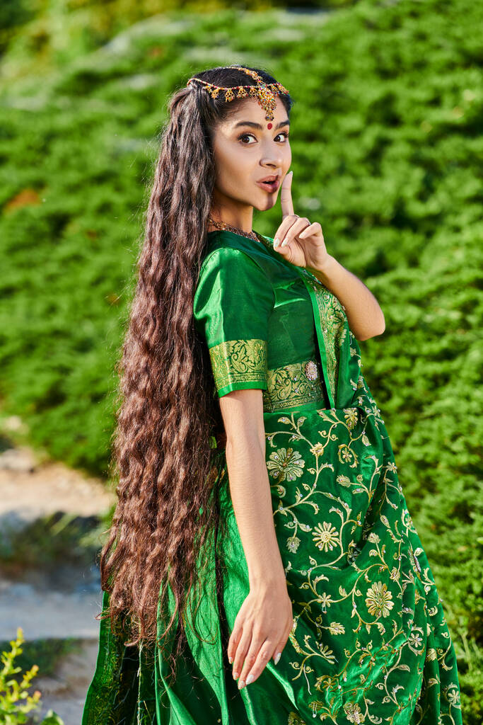 Sari 'deki genç Hintli kadın gizli jest yapıyor ve parktaki bitkilerin yakınındaki kameraya bakıyor. - Fotoğraf, Görsel